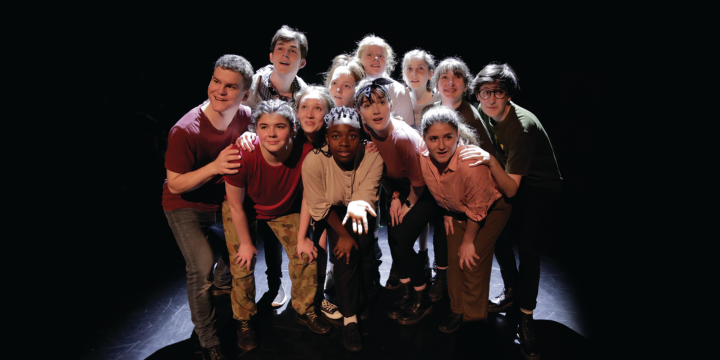 Group of young performers huddled together under a spotlight looking in different directions.